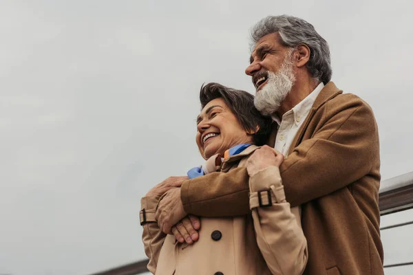 Vista de ángulo bajo de feliz hombre mayor abrazando a la esposa positiva en abrigo beige y sonriendo fuera - foto de stock