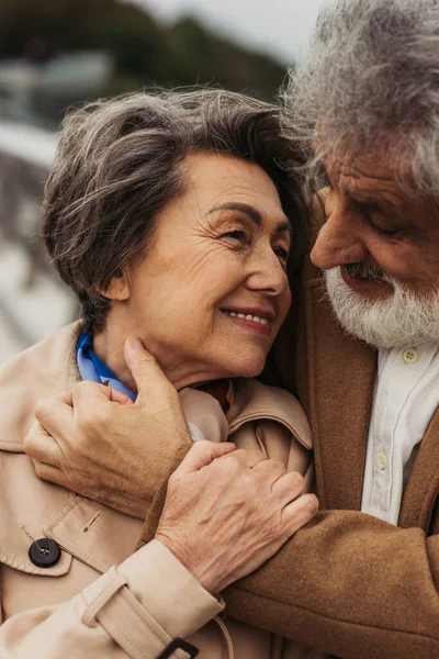 Retrato de hombre mayor feliz abrazando esposa complacida en abrigo beige y sonriendo afuera - foto de stock