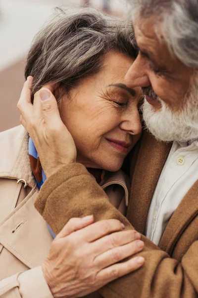 Portrait d'un homme âgé attentionné embrassant une femme heureuse en manteau beige et souriant à l'extérieur — Photo de stock