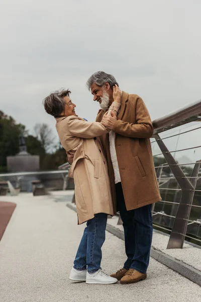 Comprimento total de mulher idosa feliz abraçando marido barbudo em casaco bege na ponte perto de trilho de guarda — Fotografia de Stock