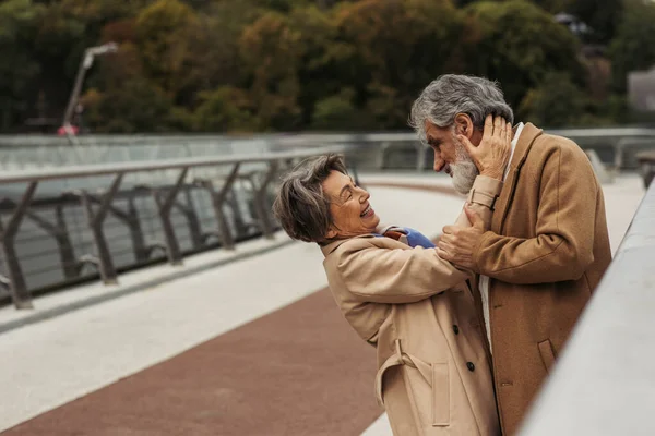 Alegre mulher idosa abraçando marido barbudo em casaco bege na ponte perto de guarda ferroviário — Fotografia de Stock