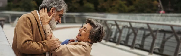 Fröhliche ältere Frau umarmt bärtigen Mann im beigen Mantel auf Brücke in der Nähe von Leitplanke, Banner — Stockfoto