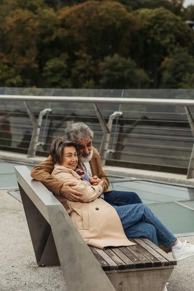Glücklicher Senior umarmt ältere Frau, während er auf Bank in der Nähe der Brückenleitplanke sitzt — Stockfoto