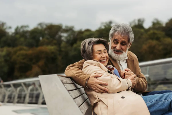 Happy senior man hugging joyful wife while sitting on bench outdoors — Stock Photo