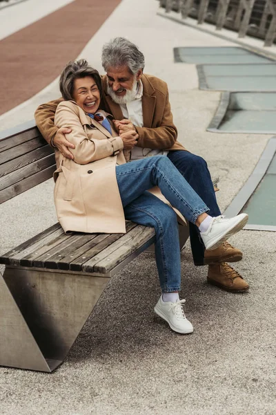 Comprimento total de feliz homem sênior abraçando alegre esposa rindo enquanto sentado no banco — Stock Photo