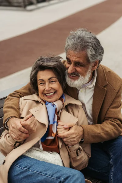 Happy senior man in coat hugging elderly wife smiling while sitting on bench — Stock Photo