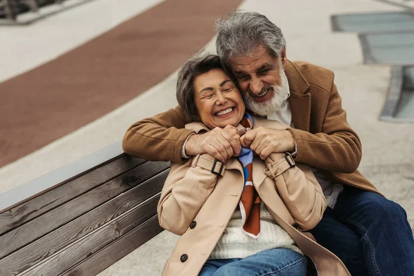 Веселый мужчина в пальто обнимает пожилую жену, улыбаясь, сидя на скамейке — стоковое фото