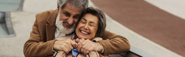 Alegre hombre mayor en abrigo abrazando anciana esposa sonriendo con los ojos cerrados - foto de stock