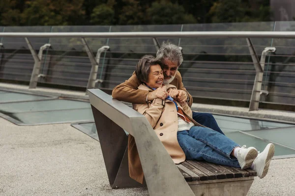 Счастливый старший мужчина обнимает позитивную жену, сидя на скамейке у ограждения моста — стоковое фото