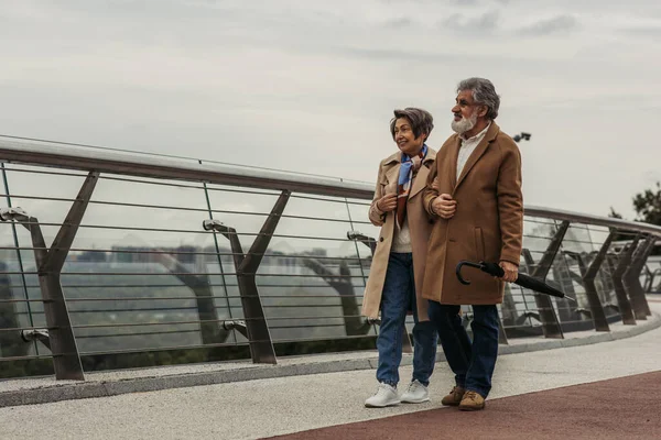Longitud completa de sonriente mujer mayor caminando con el marido barbudo sosteniendo paraguas cerca de carril de guardia del puente - foto de stock