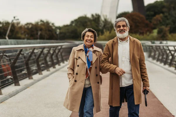 Cheerful senior woman walking with happy husband holding umbrella on bridge — Stock Photo