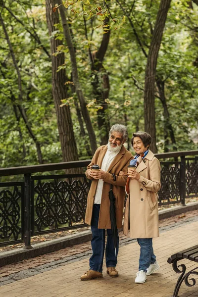 Full length of cheerful senior couple in beige coats holding paper cups and walking in park — Stock Photo