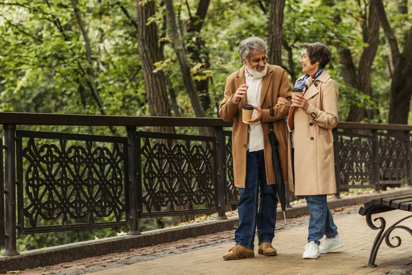 Toute la longueur du couple retraité joyeux en manteaux beige tenant des tasses en papier et marchant dans le parc — Photo de stock