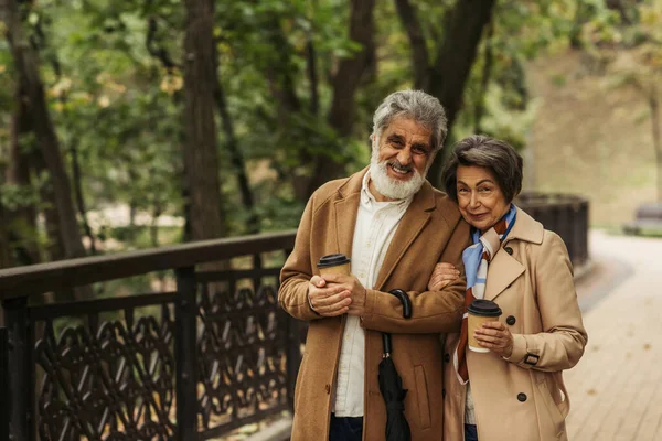 Cheerful retired couple in beige coats holding paper cups and walking in park — Stock Photo