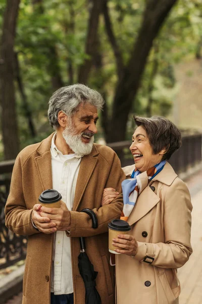 Joyful retired couple in beige coats holding paper cups and walking in park — Stock Photo