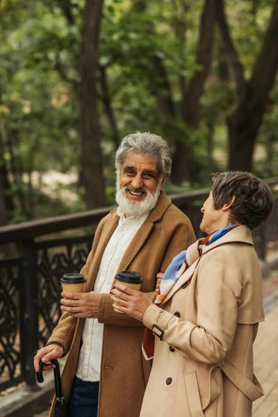 Joyeux couple retraité en manteaux beiges tenant des tasses en papier et souriant dans le parc — Photo de stock