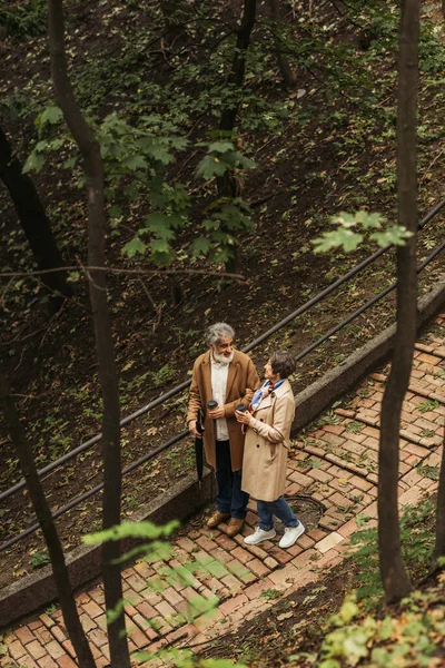 Vista ad alto angolo di gioiosa coppia in pensione in cappotti beige in possesso di bicchieri di carta e camminare nel parco — Foto stock