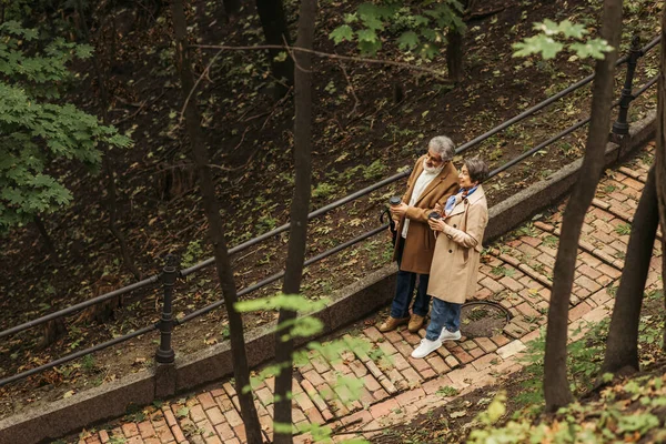 Blick aus der Vogelperspektive auf ein fröhliches Rentnerpaar in beigen Mänteln mit Pappbechern beim Spazierengehen im Park — Stockfoto