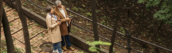 Blick aus der Vogelperspektive auf ein fröhliches Rentnerpaar in beigen Mänteln mit Pappbechern beim Spazierengehen im Park, Banner — Stockfoto