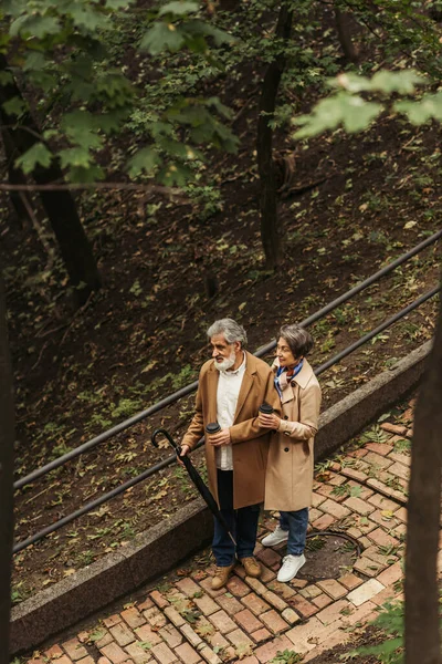High angle view of pleased and retired couple in beige coats holding paper cups while walking in park — Stock Photo