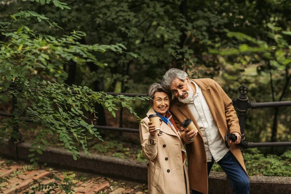 Pleased senior couple in beige coats holding paper cups in autumnal park — Stock Photo