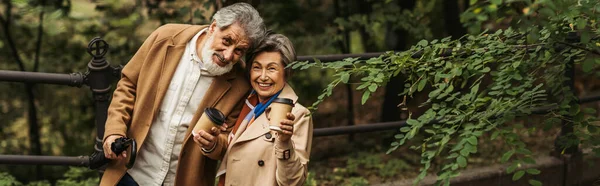 Complacida pareja de ancianos en abrigos beige sosteniendo vasos de papel en el parque otoñal, pancarta - foto de stock