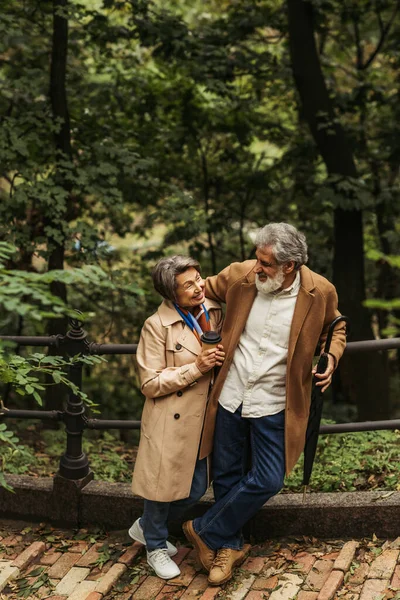 Homem sênior feliz segurando guarda-chuva e olhando para a esposa com copo de papel no parque — Stock Photo