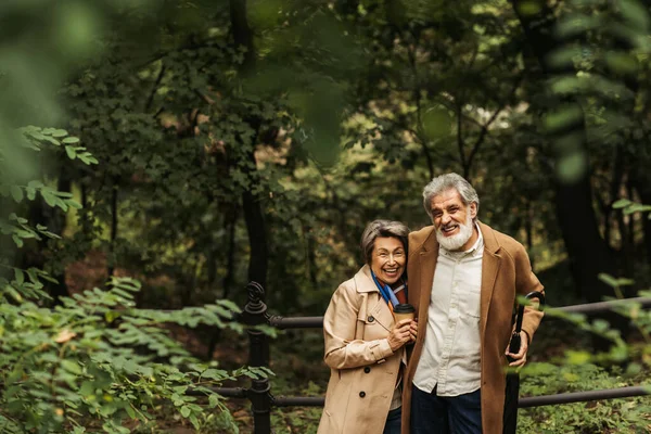 Feliz hombre mayor sosteniendo paraguas y de pie cerca de la esposa con taza de papel en el parque - foto de stock