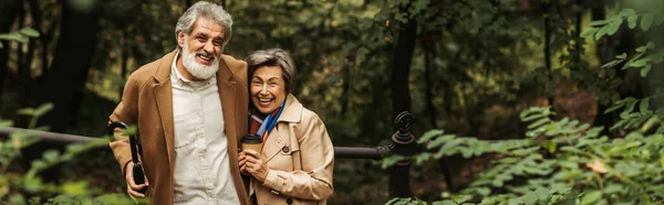 Casal sénior satisfeito em casacos bege segurando café para ir no parque outonal, banner — Fotografia de Stock