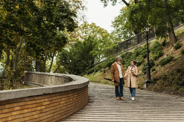 Full length view of happy senior couple in beige coats walking with coffee to go and umbrella in park — Stock Photo