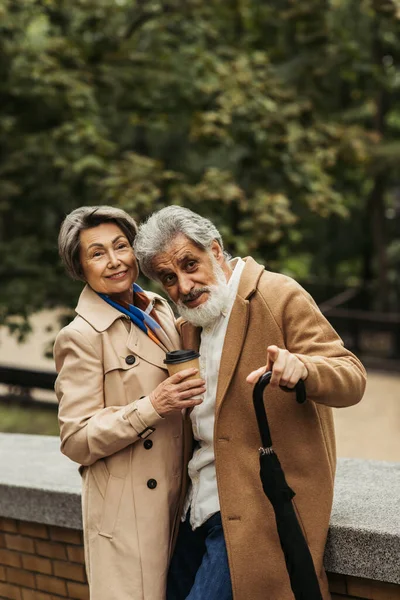 Senior hält Regenschirm und zeigt mit Finger auf Frau mit Pappbecher — Stockfoto