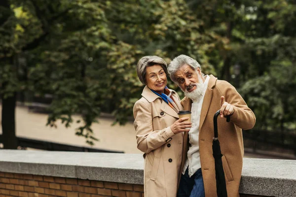 Uomo anziano che tiene l'ombrello e punta con il dito vicino alla moglie allegra con tazza di carta — Foto stock