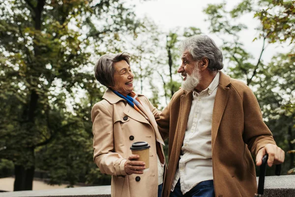 Senior hält Regenschirm in der Hand und blickt fröhliche Frau mit Pappbecher im Park an — Stockfoto