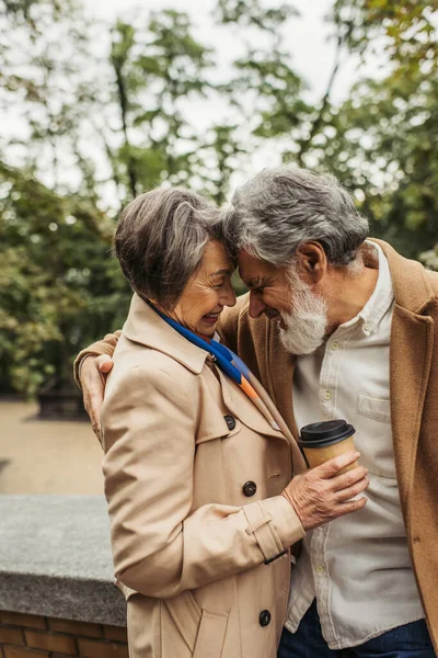 Homme âgé barbu en manteau étreignant femme gaie tenant tasse en papier avec café pour aller dans le parc — Photo de stock