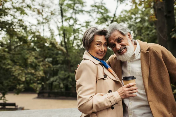 Homme âgé barbu en manteau étreignant femme heureuse tenant tasse en papier avec café pour aller dans le parc — Photo de stock