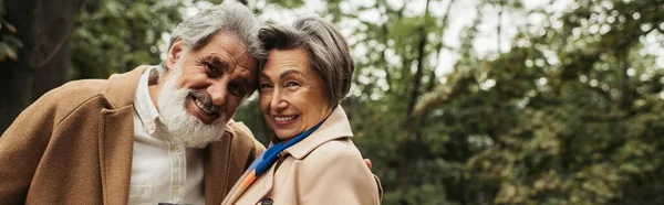 Uomo anziano barbuto in cappotto sorridente con moglie soddisfatta nel parco, striscione — Stock Photo
