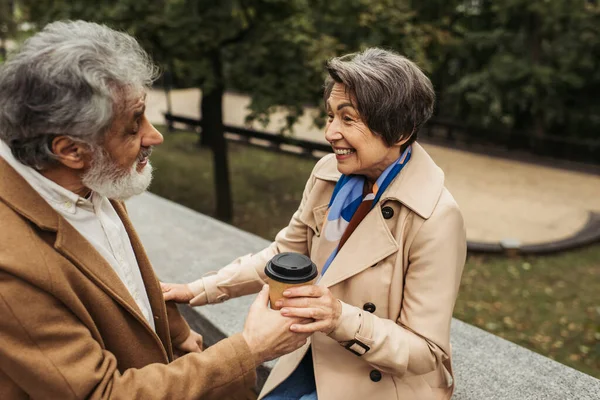 Uomo barbuto e anziano dando tazza di carta a moglie allegra in cappotto nel parco — Foto stock