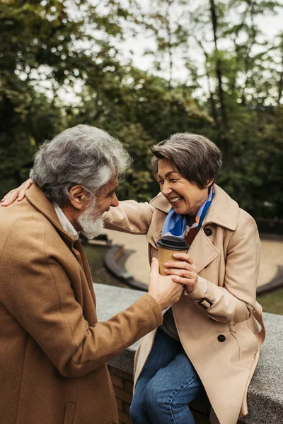 Barbudo y hombre mayor y esposa alegre en abrigo sosteniendo taza de papel en el parque - foto de stock