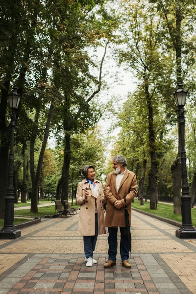 Comprimento total da mulher sênior feliz segurando copo de papel enquanto caminha com o marido elegante no casaco — Fotografia de Stock