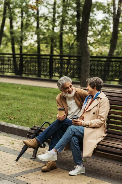 Feliz casal sênior em casacos segurando copos de papel e sentado no banco no parque — Fotografia de Stock
