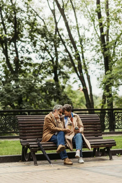 Feliz pareja jubilada en abrigos sosteniendo vasos de papel y sentado en el banco en el parque - foto de stock
