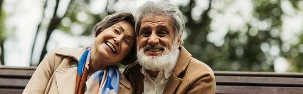 Heureux couple âgé en manteaux souriant tout en regardant la caméra dans le parc, bannière — Photo de stock