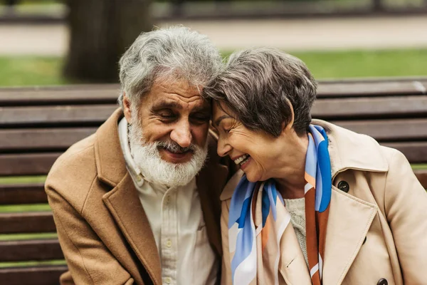 Glückliches Seniorenpaar mit geschlossenen Augen, das in Mänteln im Park sitzt und lächelt — Stockfoto