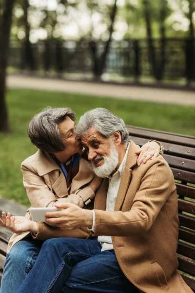 Senior femme baisers tandis que barbu mari dans manteau prise selfie dans parc — Photo de stock