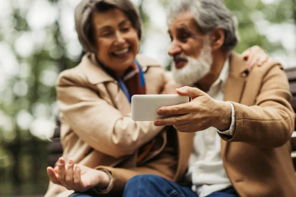 Verschwommene Seniorin lächelt, während ein bärtiger Mann im Mantel ein Selfie auf seinem Smartphone macht — Stockfoto