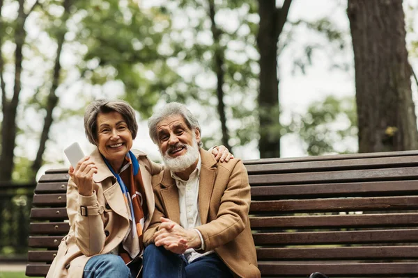 Щаслива старша жінка посміхається, тримаючи смартфон і сидячи з бородатим чоловіком в пальто на лавці — стокове фото