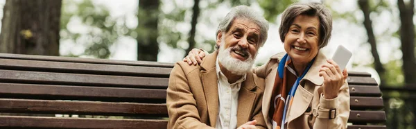 Старша жінка посміхається, тримаючи смартфон і сидячи з бородатим чоловіком в пальто на лавці, банер — стокове фото