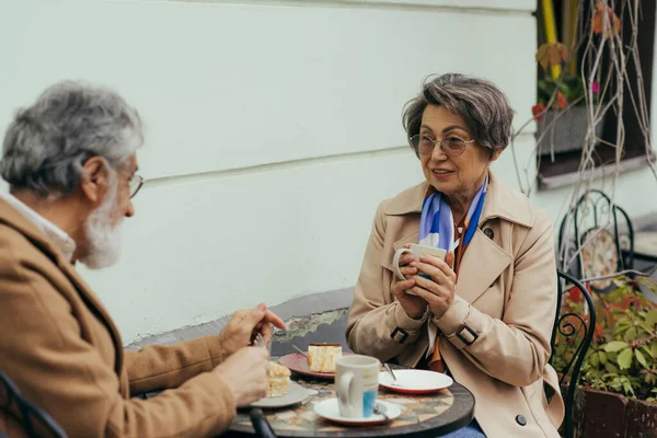 Senior woman in eyeglasses and trench coat holding cup during brunch with husband — Stock Photo