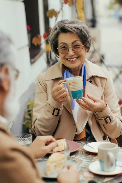 Happy senior woman in eyeglasses and trench coat holding cup during brunch with husband on terrace of cafe — Stock Photo