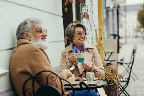 Fröhliche Seniorin in Brille und Trenchcoat beim Brunch mit Ehemann auf der Terrasse des Cafés — Stockfoto